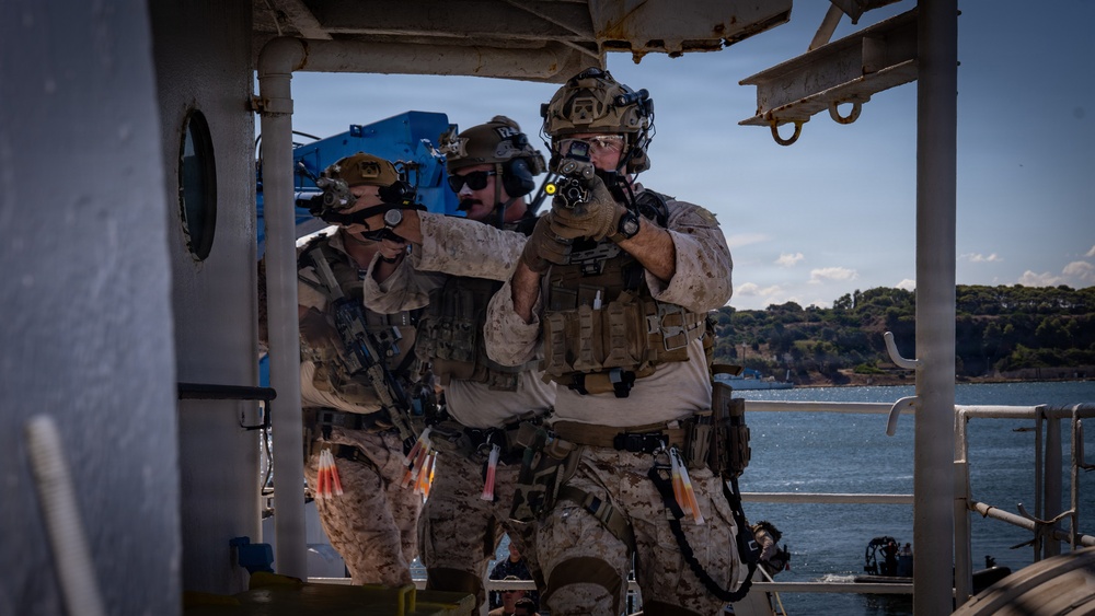 Three military personnel in desert camouflage (MARPAT) uniforms are conducting what appears to be a maritime vessel boarding or ship-clearing exercise on the deck of a vessel. The operators are equipped with full tactical gear including helmets with night vision device mounts, plate carriers, and are aiming firearms in a coordinated formation. The background shows a coastal waterway with tree-covered hills, suggesting a Mediterranean or similar temperate coastal environment.