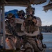 Three military personnel in desert camouflage (MARPAT) uniforms are conducting what appears to be a maritime vessel boarding or ship-clearing exercise on the deck of a vessel. The operators are equipped with full tactical gear including helmets with night vision device mounts, plate carriers, and are aiming firearms in a coordinated formation. The background shows a coastal waterway with tree-covered hills, suggesting a Mediterranean or similar temperate coastal environment.