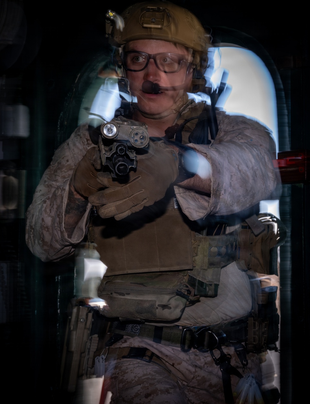 A military or law enforcement operator in full tactical gear is photographed through what appears to be a glass or reflective surface, creating a double-exposure or reflection effect. The individual is holding a pistol with a mounted weapon light in a two-handed grip, pointed toward the camera. The image has a dark background with an illuminated doorway or hatch visible behind the subject.
