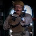 A military or law enforcement operator in full tactical gear is photographed through what appears to be a glass or reflective surface, creating a double-exposure or reflection effect. The individual is holding a pistol with a mounted weapon light in a two-handed grip, pointed toward the camera. The image has a dark background with an illuminated doorway or hatch visible behind the subject.