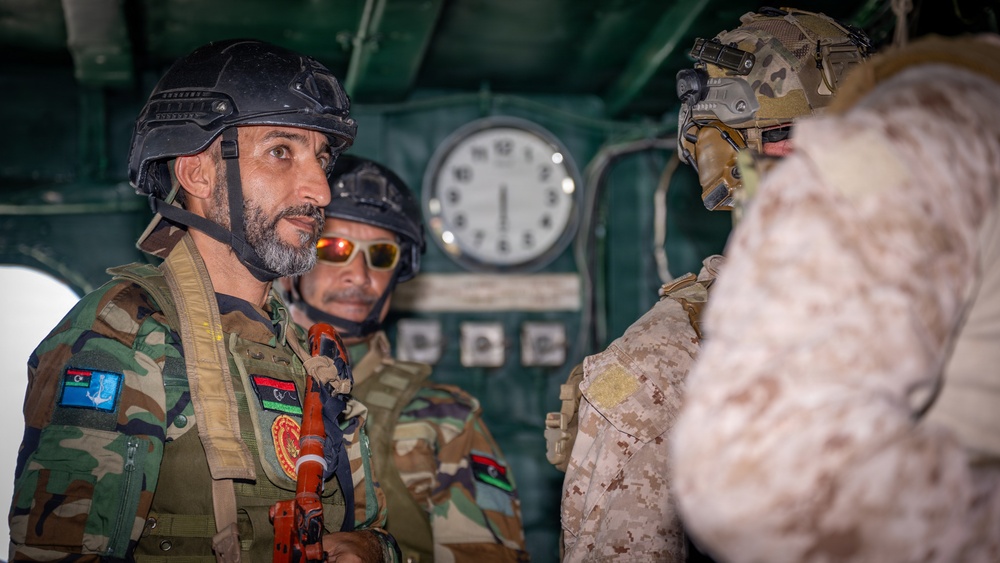 The image shows military personnel in tactical gear engaged in conversation, likely aboard a naval vessel or military transport given the interior setting with a clock and control panels visible in the background. At least two individuals are wearing patches consistent with the Libyan flag and insignia, suggesting they are members of Libyan armed forces. A third individual in desert camouflage pattern gear faces them, indicating a multi-party military interaction or briefing.