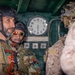 The image shows military personnel in tactical gear engaged in conversation, likely aboard a naval vessel or military transport given the interior setting with a clock and control panels visible in the background. At least two individuals are wearing patches consistent with the Libyan flag and insignia, suggesting they are members of Libyan armed forces. A third individual in desert camouflage pattern gear faces them, indicating a multi-party military interaction or briefing.