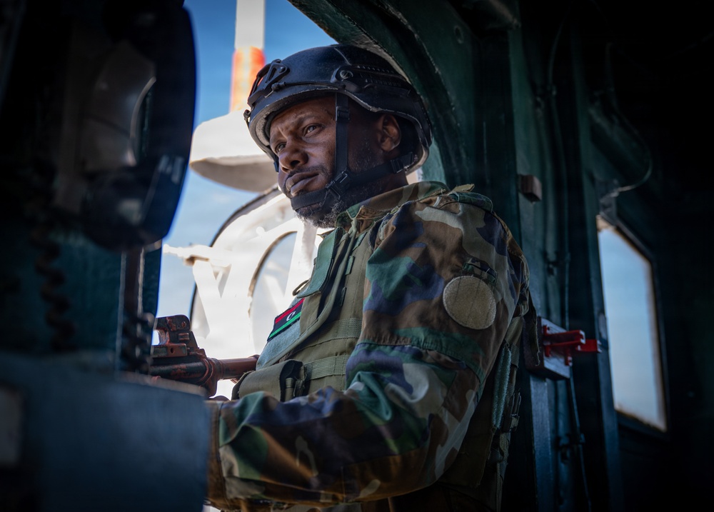 A uniformed military soldier is positioned inside what appears to be a military vehicle or aircraft doorway. He is wearing camouflage fatigues with a flag patch on the sleeve, a tactical helmet, and appears to be manning or operating equipment. The image is dramatically lit with a bright background contrasting against the darker interior.