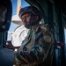 A uniformed military soldier is positioned inside what appears to be a military vehicle or aircraft doorway. He is wearing camouflage fatigues with a flag patch on the sleeve, a tactical helmet, and appears to be manning or operating equipment. The image is dramatically lit with a bright background contrasting against the darker interior.