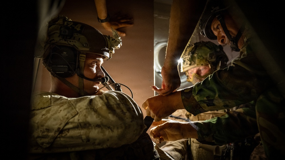 Military personnel are engaged in what appears to be a tactical medical or casualty care scenario, likely inside a military aircraft or vehicle. One soldier wearing a tactical helmet with communications equipment is being attended to by at least one other service member. The scene is dramatically lit with low ambient light, suggesting a nighttime or low-light operational environment.