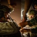 Military personnel are engaged in what appears to be a tactical medical or casualty care scenario, likely inside a military aircraft or vehicle. One soldier wearing a tactical helmet with communications equipment is being attended to by at least one other service member. The scene is dramatically lit with low ambient light, suggesting a nighttime or low-light operational environment.