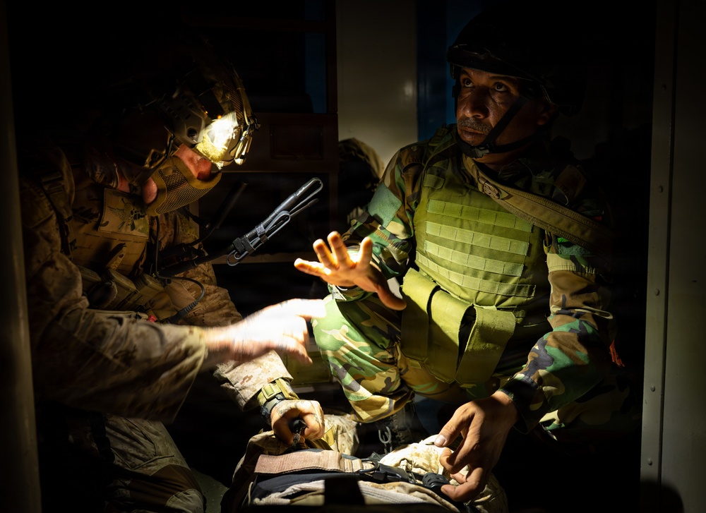 Two military personnel are engaged in what appears to be a tactical briefing or equipment check in a low-light environment. One soldier wearing a helmet with a mounted light source is illuminating a bag or kit, while a second soldier in green camouflage with a plate carrier vest gestures with an open hand. The scene suggests a training exercise or operational preparation scenario.