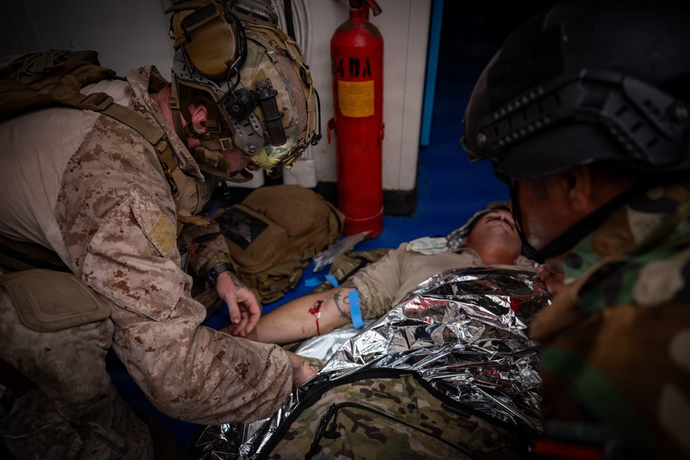 Two military personnel in combat gear are providing tactical combat casualty care (TCCC) to a downed soldier lying on a blue surface. The casualty is wrapped in a mylar emergency blanket and has visible simulated or actual blood on their arm. A red fire extinguisher is visible in the background, suggesting an indoor or enclosed environment such as a ship or building.