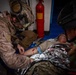 Two military personnel in combat gear are providing tactical combat casualty care (TCCC) to a downed soldier lying on a blue surface. The casualty is wrapped in a mylar emergency blanket and has visible simulated or actual blood on their arm. A red fire extinguisher is visible in the background, suggesting an indoor or enclosed environment such as a ship or building.