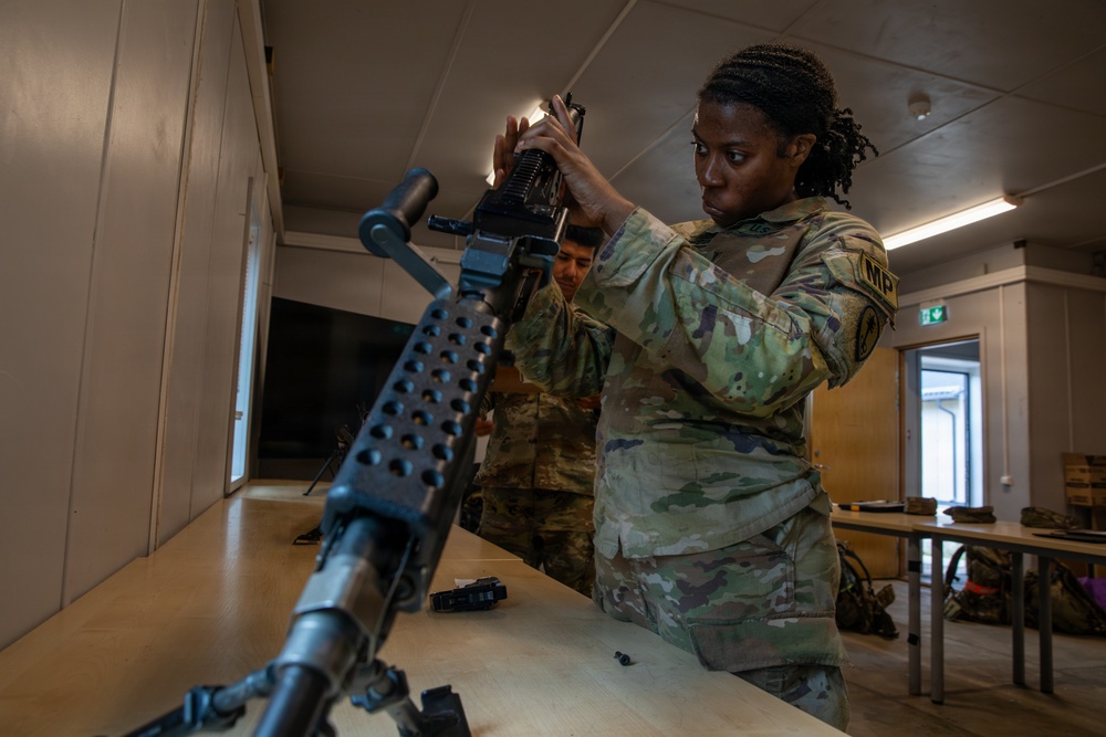 Two U.S. Army soldiers in Operational Camouflage Pattern (OCP) uniforms are working with military weapons on a table in an indoor facility. The soldier in the foreground, wearing an 'MP' (Military Police) patch, is handling what appears to be an M249 or similar light machine gun. The setting appears to be a training or maintenance environment where weapons assembly or disassembly is being practiced.