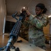 Two U.S. Army soldiers in Operational Camouflage Pattern (OCP) uniforms are working with military weapons on a table in an indoor facility. The soldier in the foreground, wearing an 'MP' (Military Police) patch, is handling what appears to be an M249 or similar light machine gun. The setting appears to be a training or maintenance environment where weapons assembly or disassembly is being practiced.