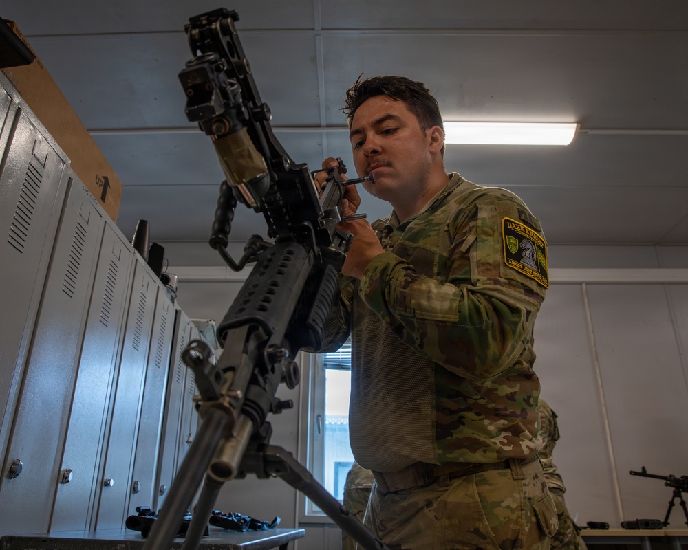 A soldier in MultiCam/OCP camouflage uniform is performing maintenance or assembly on a crew-served machine gun, likely an M240 or similar weapon system, mounted on a bipod/tripod in an indoor armory or maintenance room. The soldier has a tool or small part held in his mouth while working with both hands on the weapon's upper receiver area. A unit patch reading 'Dark Knights' is visible on his left shoulder.