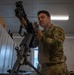 A soldier in MultiCam/OCP camouflage uniform is performing maintenance or assembly on a crew-served machine gun, likely an M240 or similar weapon system, mounted on a bipod/tripod in an indoor armory or maintenance room. The soldier has a tool or small part held in his mouth while working with both hands on the weapon's upper receiver area. A unit patch reading 'Dark Knights' is visible on his left shoulder.