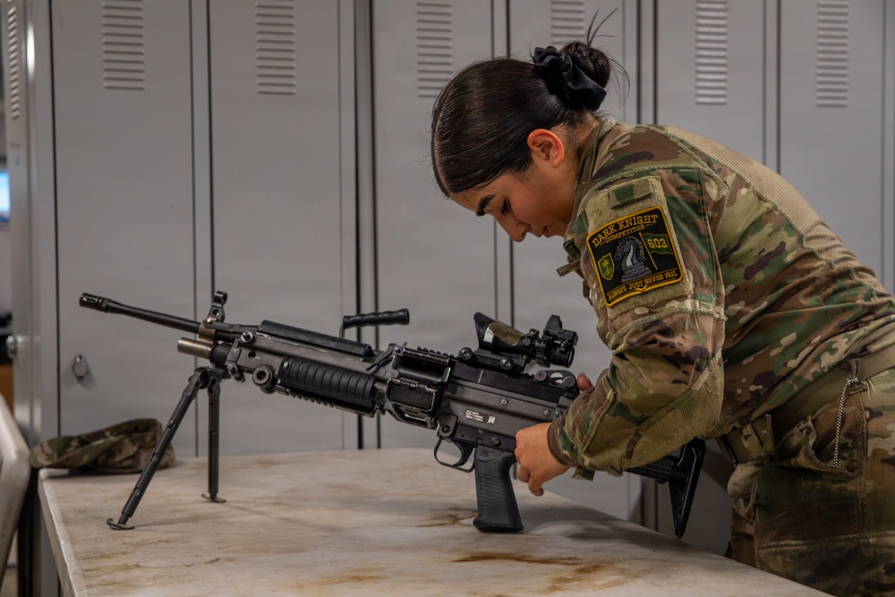 A female soldier in MultiCam/OCP camouflage uniform is inspecting or handling a light machine gun on a table in what appears to be an armory or equipment room. She is wearing a patch on her right shoulder that reads 'Dark Knight Competitor' with the number 603. The setting includes gray metal lockers in the background, suggesting a military facility.