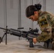 A female soldier in MultiCam/OCP camouflage uniform is inspecting or handling a light machine gun on a table in what appears to be an armory or equipment room. She is wearing a patch on her right shoulder that reads 'Dark Knight Competitor' with the number 603. The setting includes gray metal lockers in the background, suggesting a military facility.