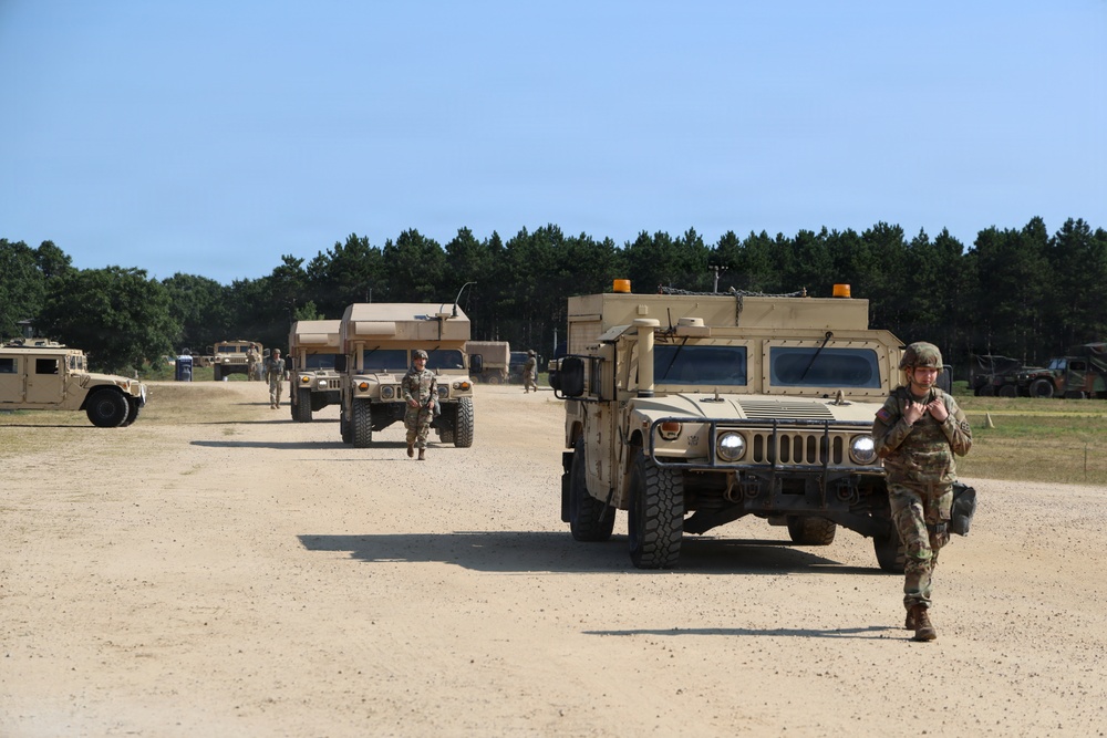 The image shows a military convoy or staging area with multiple tan/desert-colored HMMWV (Humvee) vehicles lined up on a sandy dirt road. Several soldiers in MultiCam/OCP camouflage uniforms are visible walking alongside and between the vehicles. The setting appears to be a training installation or forward operating base, with a treeline of pine trees visible in the background.