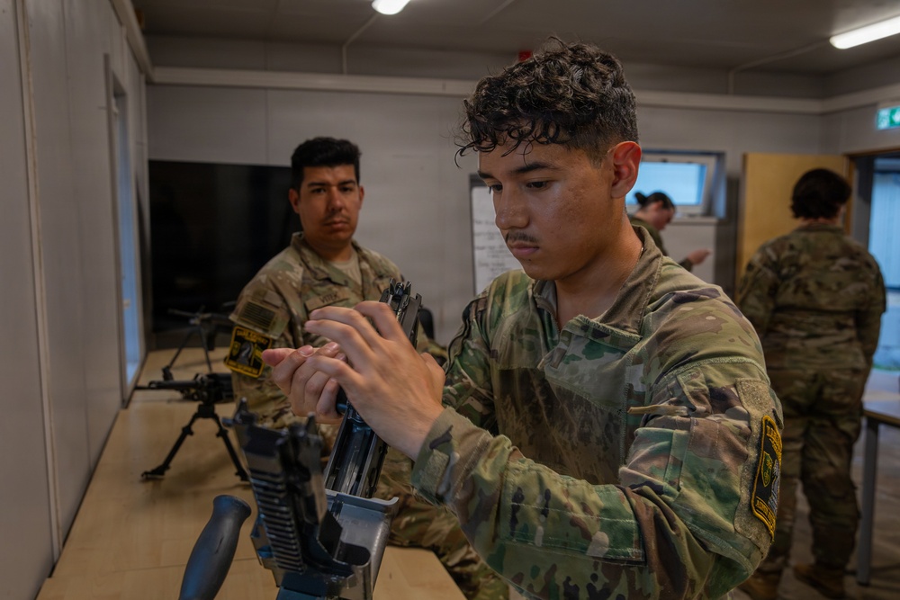 Military personnel in MultiCam/OCP camouflage uniforms are engaged in a weapons maintenance or familiarization training session indoors. The foreground soldier is handling and inspecting what appears to be a disassembled machine gun on a table, while others observe or conduct similar activities in the background. The setting appears to be a training facility or classroom environment.