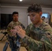Military personnel in MultiCam/OCP camouflage uniforms are engaged in a weapons maintenance or familiarization training session indoors. The foreground soldier is handling and inspecting what appears to be a disassembled machine gun on a table, while others observe or conduct similar activities in the background. The setting appears to be a training facility or classroom environment.