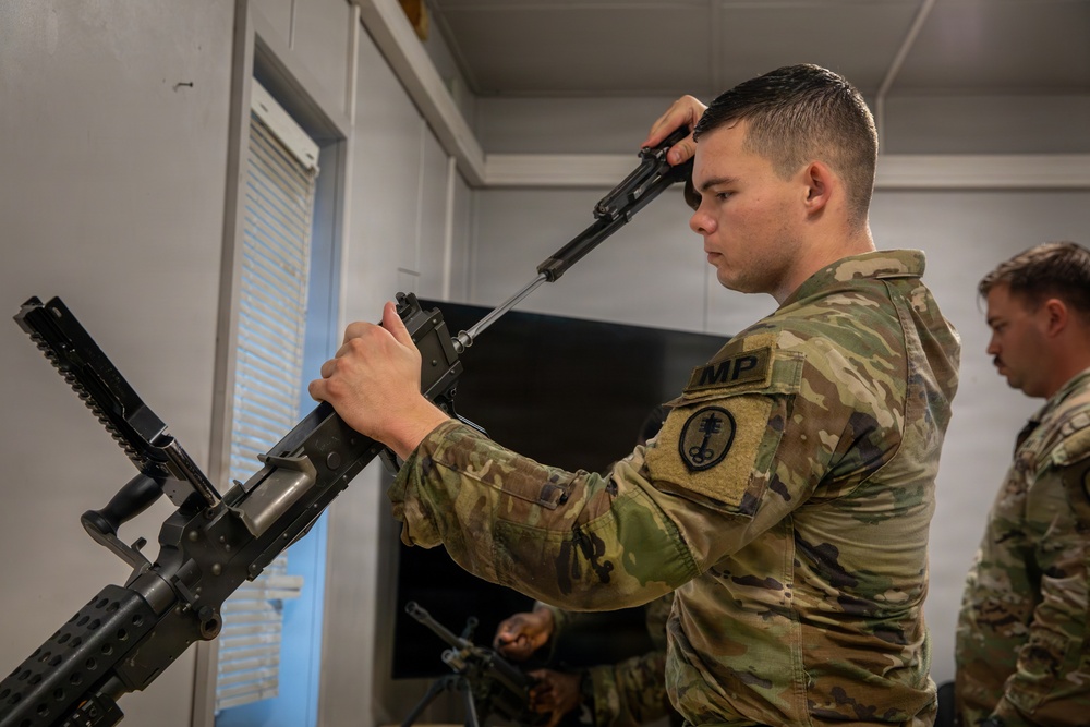 A U.S. Army Military Police (MP) soldier is performing a field strip or maintenance procedure on what appears to be an M249 Squad Automatic Weapon (SAW) or similar belt-fed machine gun in an indoor training environment. The soldier is holding a component of the weapon up near his head, likely inspecting or reassembling the bolt carrier or feed tray cover. A second soldier is visible in the background, also engaged with a weapon.