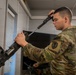 A U.S. Army Military Police (MP) soldier is performing a field strip or maintenance procedure on what appears to be an M249 Squad Automatic Weapon (SAW) or similar belt-fed machine gun in an indoor training environment. The soldier is holding a component of the weapon up near his head, likely inspecting or reassembling the bolt carrier or feed tray cover. A second soldier is visible in the background, also engaged with a weapon.