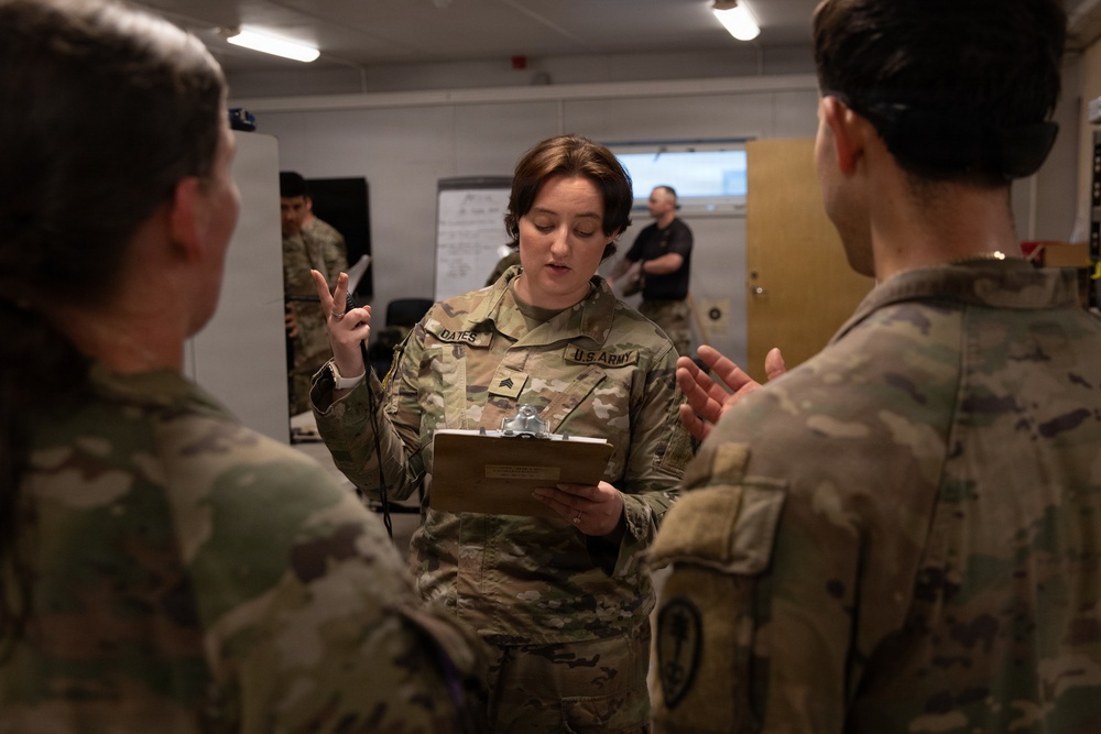 A U.S. Army soldier with the name tape 'OATES' and a Staff Sergeant rank insignia is holding a clipboard and briefing or instructing two other soldiers in what appears to be an indoor military operations or briefing room. She is gesturing with her hand while speaking, suggesting she is explaining or coordinating a task. Additional personnel are visible in the background near a whiteboard.