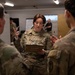 A U.S. Army soldier with the name tape 'OATES' and a Staff Sergeant rank insignia is holding a clipboard and briefing or instructing two other soldiers in what appears to be an indoor military operations or briefing room. She is gesturing with her hand while speaking, suggesting she is explaining or coordinating a task. Additional personnel are visible in the background near a whiteboard.