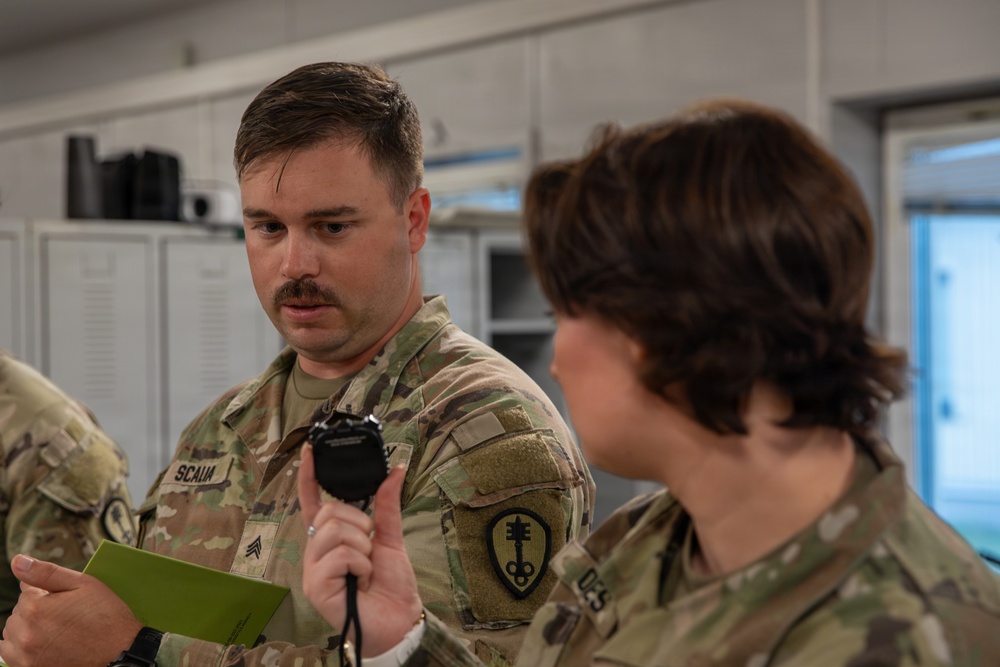 Two U.S. Army soldiers in OCP (Operational Camouflage Pattern) uniforms are engaged in what appears to be a training or briefing activity indoors. The soldier in focus, identified by the name tape 'SCALIA' and wearing Sergeant rank insignia, is holding a stopwatch and referencing a small green notepad. A unit patch consistent with a Cyber or Signal branch is visible on his left shoulder.