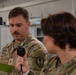 Two U.S. Army soldiers in OCP (Operational Camouflage Pattern) uniforms are engaged in what appears to be a training or briefing activity indoors. The soldier in focus, identified by the name tape 'SCALIA' and wearing Sergeant rank insignia, is holding a stopwatch and referencing a small green notepad. A unit patch consistent with a Cyber or Signal branch is visible on his left shoulder.