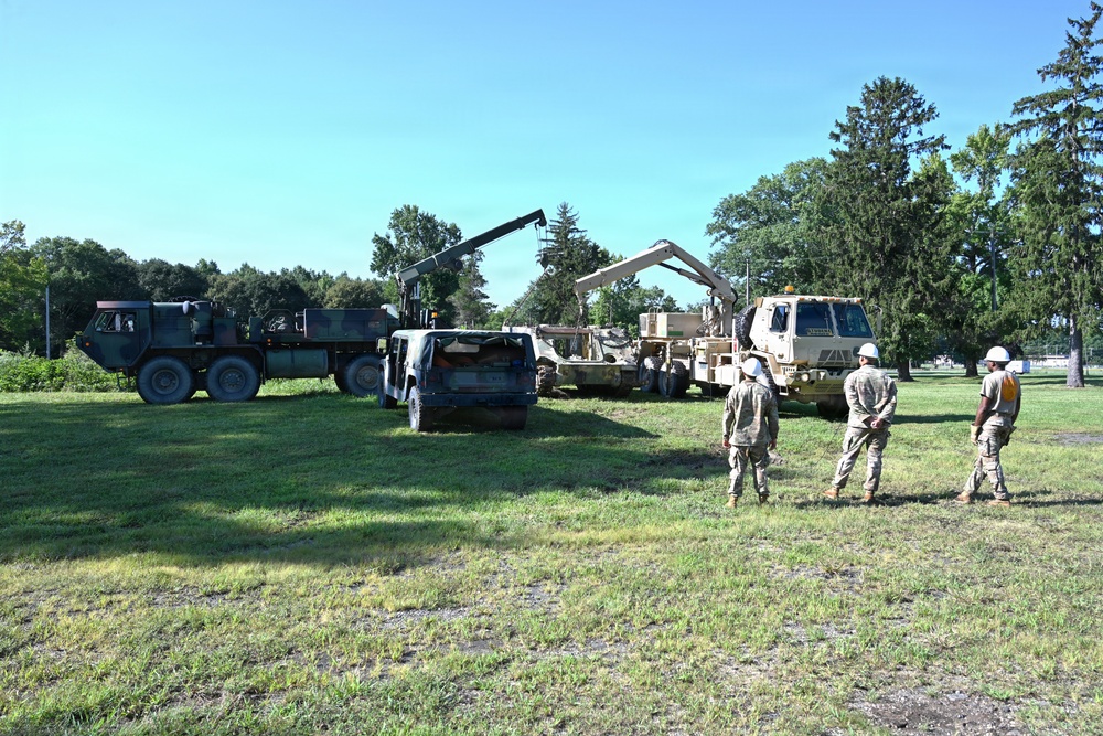 Military personnel are conducting a vehicle recovery or lifting operation in an open grassy field, likely on a military installation. Two large military trucks equipped with crane/boom arms are being used to lift or move what appears to be a disabled or damaged civilian vehicle. Three soldiers wearing helmets and military uniforms are observing and supervising the operation.