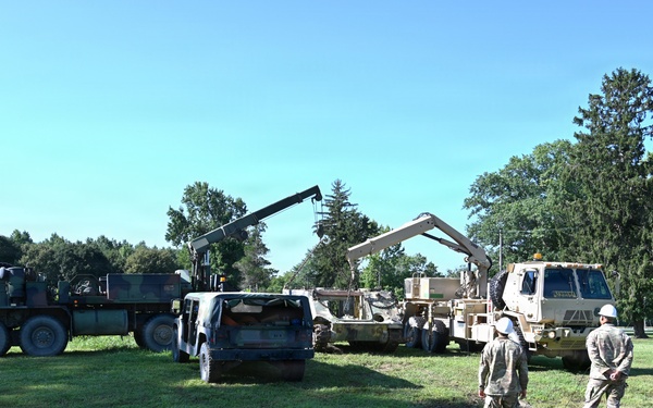 Military personnel are conducting a vehicle recovery or lifting operation in an open grassy field, likely on a military installation. Two large military trucks equipped with crane/boom arms are being used to lift or move what appears to be a disabled or damaged civilian vehicle. Three soldiers wearing helmets and military uniforms are observing and supervising the operation.