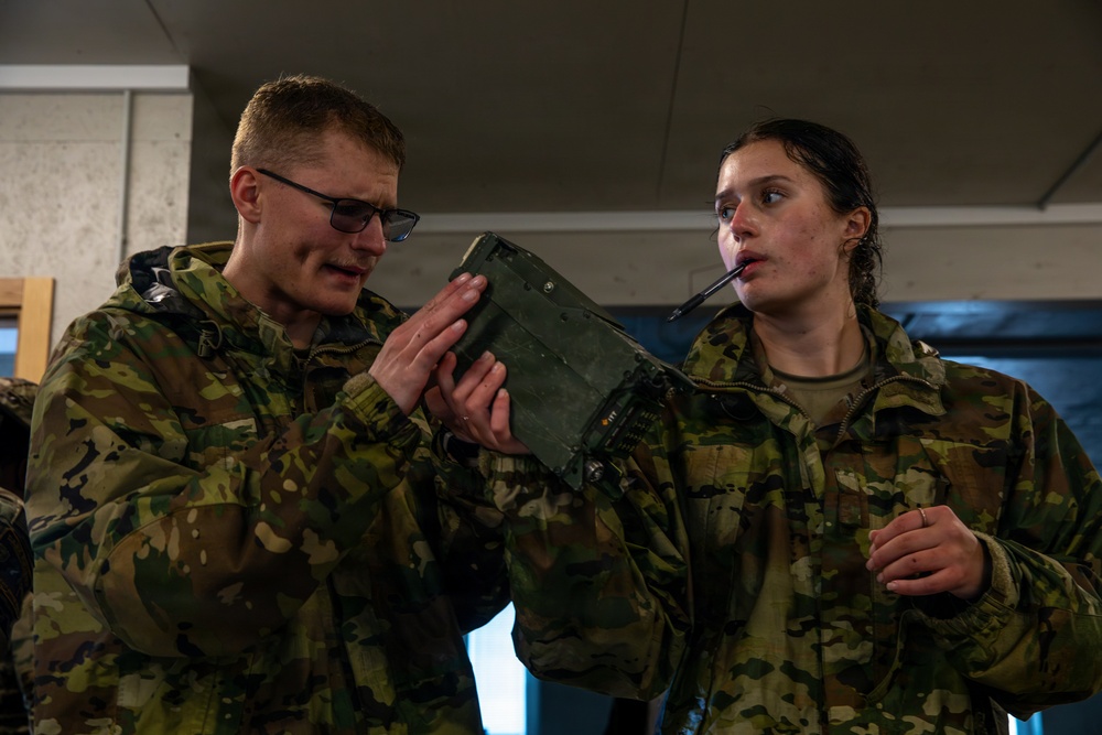 Two military personnel in camouflage uniforms are examining a military field radio or communication device together indoors. The male soldier on the left is holding and inspecting the olive-drab colored equipment while the female soldier on the right observes closely. Both appear wet, suggesting recent outdoor activity in wet conditions.