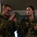Two military personnel in camouflage uniforms are examining a military field radio or communication device together indoors. The male soldier on the left is holding and inspecting the olive-drab colored equipment while the female soldier on the right observes closely. Both appear wet, suggesting recent outdoor activity in wet conditions.