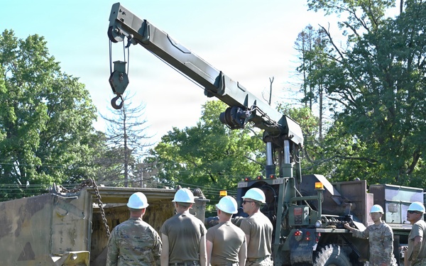 A group of military personnel in camouflage uniforms and white hard hats are gathered around military vehicles during what appears to be a recovery or maintenance training exercise. A crane-equipped heavy military truck is positioned next to a tracked armored vehicle, suggesting a vehicle recovery operation. The setting is an open grassy area surrounded by trees under clear daylight conditions.