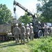 A group of military personnel in camouflage uniforms and white hard hats are gathered around military vehicles during what appears to be a recovery or maintenance training exercise. A crane-equipped heavy military truck is positioned next to a tracked armored vehicle, suggesting a vehicle recovery operation. The setting is an open grassy area surrounded by trees under clear daylight conditions.