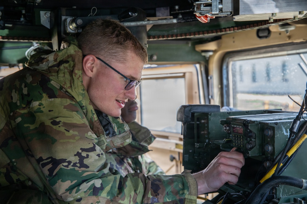 A young soldier in MultiCam/OCP camouflage uniform is operating radio communications equipment inside a military vehicle, likely a HMMWV (Humvee). He is wearing military-issue glasses and is focused on adjusting controls on a mounted tactical radio unit. A second soldier is partially visible in the background.
