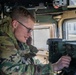 A young soldier in MultiCam/OCP camouflage uniform is operating radio communications equipment inside a military vehicle, likely a HMMWV (Humvee). He is wearing military-issue glasses and is focused on adjusting controls on a mounted tactical radio unit. A second soldier is partially visible in the background.