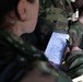 A person in military camouflage uniform is holding a small spiral-bound notepad containing a handwritten numbered list, likely operational or procedural notes. A second person in camouflage is visible in the background. The image appears to be taken in a field or tactical environment.