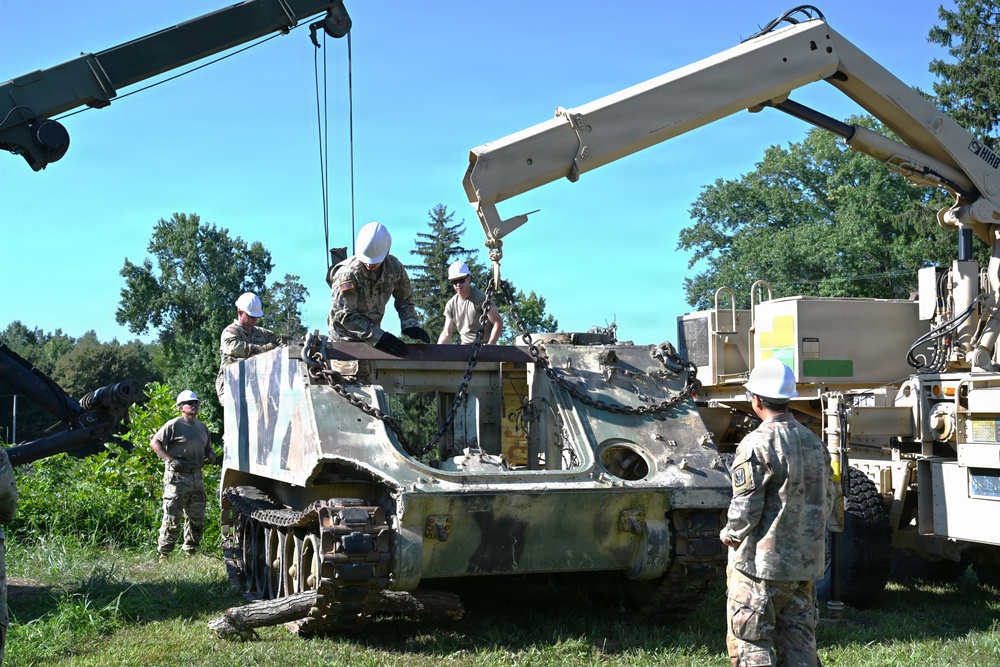 Military personnel in camouflage uniforms and white hard hats are using two crane vehicles to lift or recover a heavily damaged tracked armored vehicle, likely an M113 armored personnel carrier. The operation appears to be a recovery or salvage exercise taking place in a grassy outdoor area surrounded by trees. At least four soldiers are actively involved, with some on top of the damaged vehicle attaching chains while others observe and supervise.