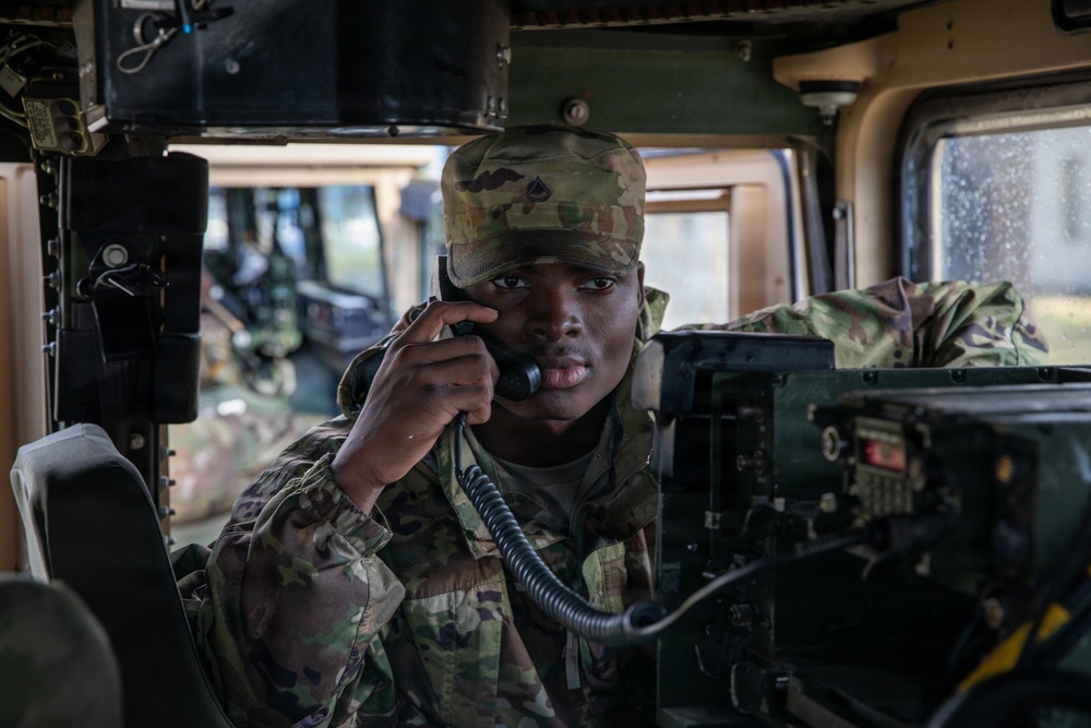 A U.S. Army soldier in Operational Camouflage Pattern (OCP) uniform is seated inside a military vehicle, likely a HMMWV (Humvee), using a radio handset to communicate. The soldier wears a patrol cap with a visible rank insignia and is surrounded by military communications equipment. The image appears to be taken during a training exercise or operational deployment.