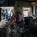 A U.S. Army soldier in Operational Camouflage Pattern (OCP) uniform is seated inside a military vehicle, likely a HMMWV (Humvee), using a radio handset to communicate. The soldier wears a patrol cap with a visible rank insignia and is surrounded by military communications equipment. The image appears to be taken during a training exercise or operational deployment.