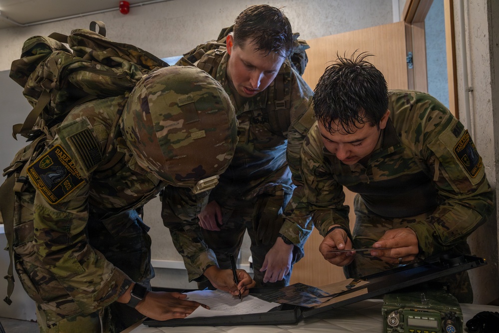 Three military personnel in MultiCam/OCP camouflage uniforms are gathered around a table, reviewing and writing on documents. Their hair appears wet, suggesting recent physical activity or exposure to weather. A radio/communications device is visible on the table alongside what appears to be a map or briefing materials.