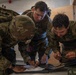 Three military personnel in MultiCam/OCP camouflage uniforms are gathered around a table, reviewing and writing on documents. Their hair appears wet, suggesting recent physical activity or exposure to weather. A radio/communications device is visible on the table alongside what appears to be a map or briefing materials.