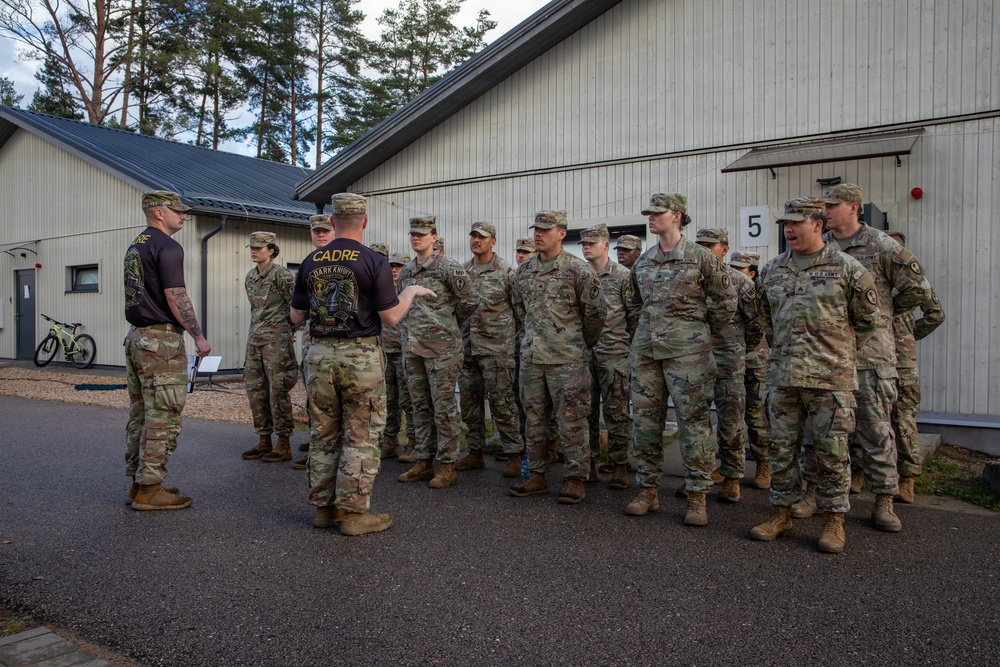 A group of U.S. Army soldiers in OCP (Operational Camouflage Pattern) uniforms are being addressed by two cadre members wearing black 'CADRE - Dark Knights' t-shirts outside a military facility. The cadre appear to be conducting a briefing or formation, likely at a training installation. The setting appears to be a European military base, suggested by the architectural style of the buildings.