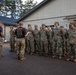 A group of U.S. Army soldiers in OCP (Operational Camouflage Pattern) uniforms are being addressed by two cadre members wearing black 'CADRE - Dark Knights' t-shirts outside a military facility. The cadre appear to be conducting a briefing or formation, likely at a training installation. The setting appears to be a European military base, suggested by the architectural style of the buildings.