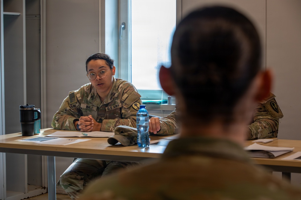 Two U.S. Army soldiers in OCP (Operational Camouflage Pattern) uniforms are seated across from each other at a table in what appears to be a military facility or office setting. The soldier facing the camera, identified by name tape as 'HALE,' appears to be engaged in a briefing, counseling session, or interview. Documents, a water bottle, and a tumbler are visible on the table between them.