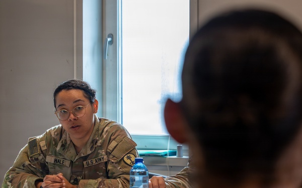 Two U.S. Army soldiers in OCP (Operational Camouflage Pattern) uniforms are seated across from each other at a table in what appears to be a military facility or office setting. The soldier facing the camera, identified by name tape as 'HALE,' appears to be engaged in a briefing, counseling session, or interview. Documents, a water bottle, and a tumbler are visible on the table between them.
