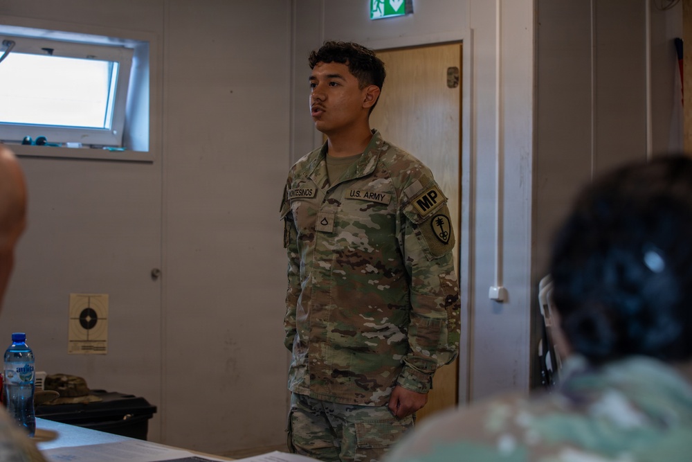 A U.S. Army Military Police (MP) soldier, identified by name tape as 'Montesinos,' is standing and speaking in what appears to be a briefing or classroom setting. The soldier is wearing Operational Camouflage Pattern (OCP) uniform with U.S. Army and MP patches visible on the left sleeve. The setting appears to be an indoor military facility with other personnel seated in the foreground.