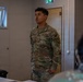 A U.S. Army Military Police (MP) soldier, identified by name tape as 'Montesinos,' is standing and speaking in what appears to be a briefing or classroom setting. The soldier is wearing Operational Camouflage Pattern (OCP) uniform with U.S. Army and MP patches visible on the left sleeve. The setting appears to be an indoor military facility with other personnel seated in the foreground.