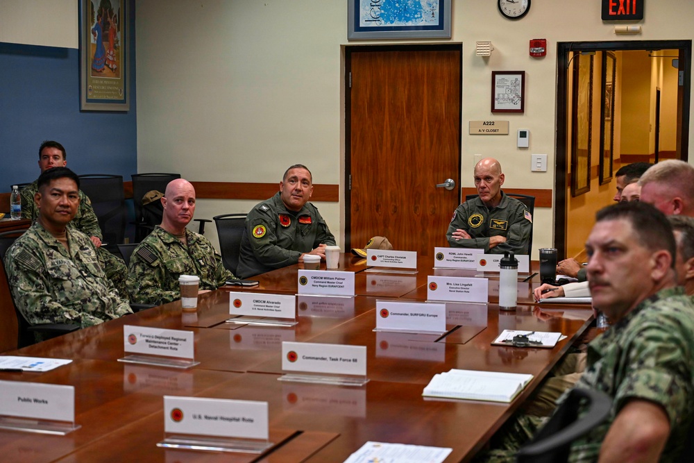 A formal military briefing or meeting is taking place in a conference room, with multiple uniformed personnel seated around a large wooden table. Participants wear various U.S. military uniforms including Navy digital camouflage, Marine Corps MARPAT, and olive green flight suits. Name placards are visible on the table identifying positions and commands.