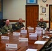 A formal military briefing or meeting is taking place in a conference room, with multiple uniformed personnel seated around a large wooden table. Participants wear various U.S. military uniforms including Navy digital camouflage, Marine Corps MARPAT, and olive green flight suits. Name placards are visible on the table identifying positions and commands.